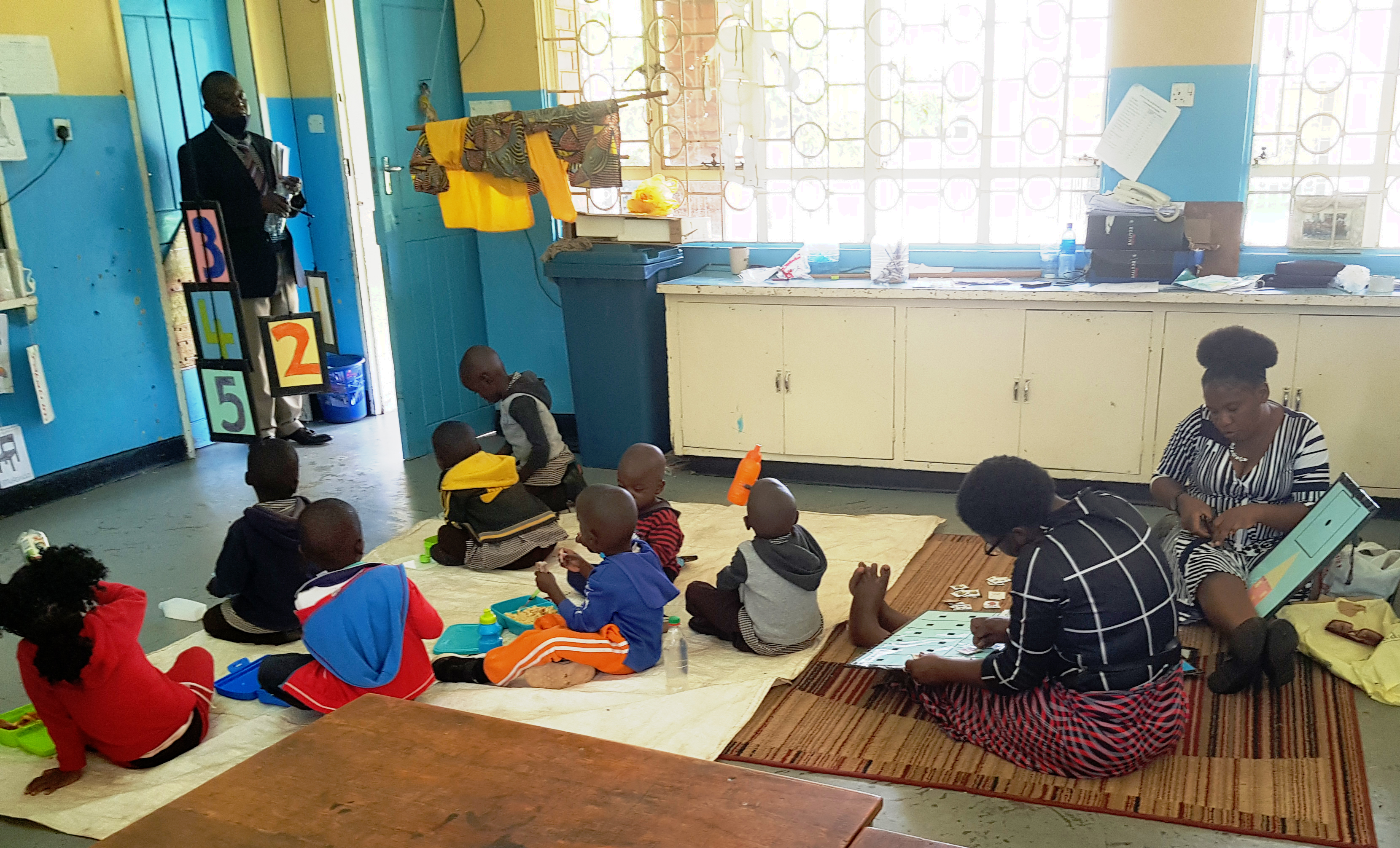 "Young students in a classroom in Zambia sitting on the floor. There are two techers sitting behind the children working and an educator standing at the door at the front of the room"