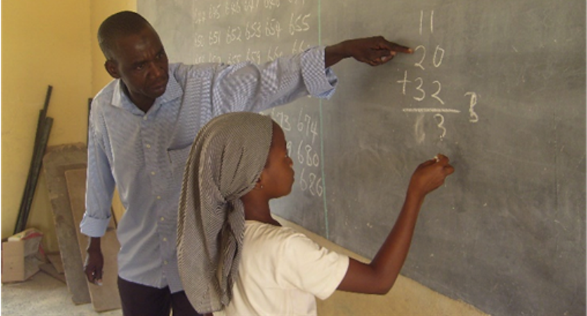 "Teacher working with a student on a math problem in front of a chalk board in a classroom in Nigeria"