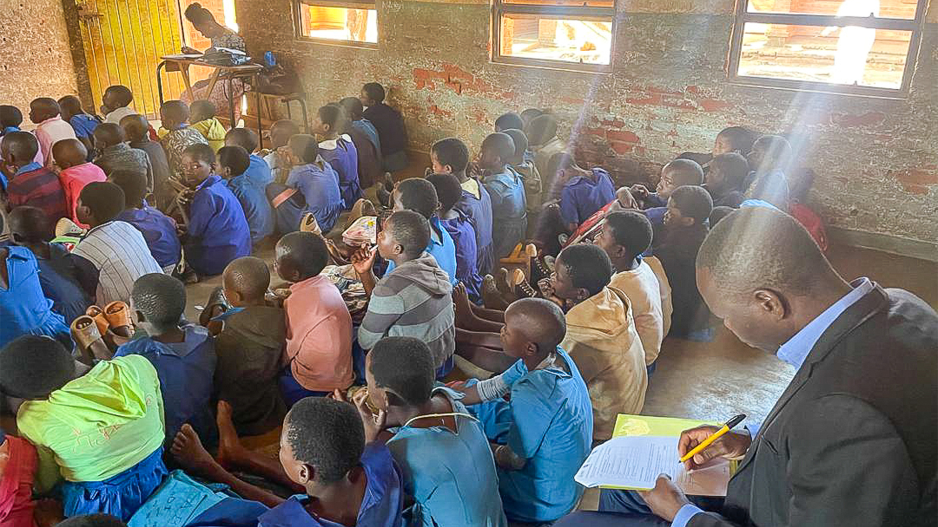 "Man sitting in the back of a classroom in Malawi taking notes on the instruction of the children."