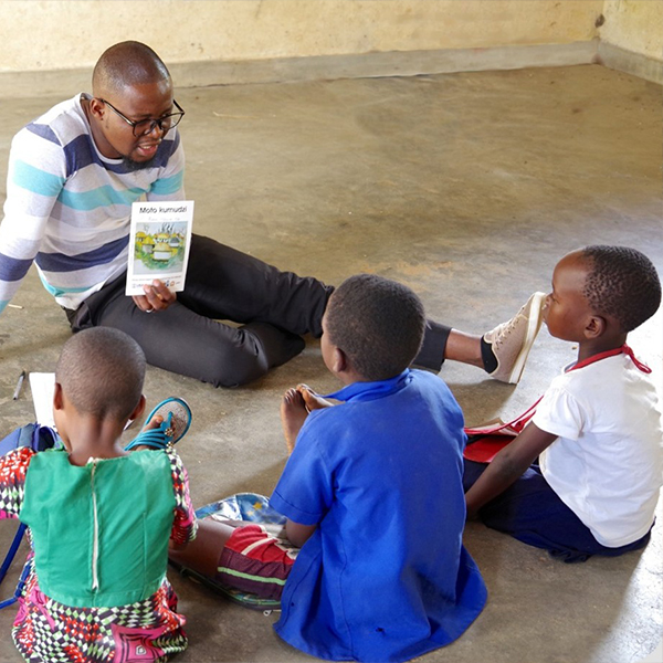 Teacher holding a book with young students sitting next to him on the floor.
