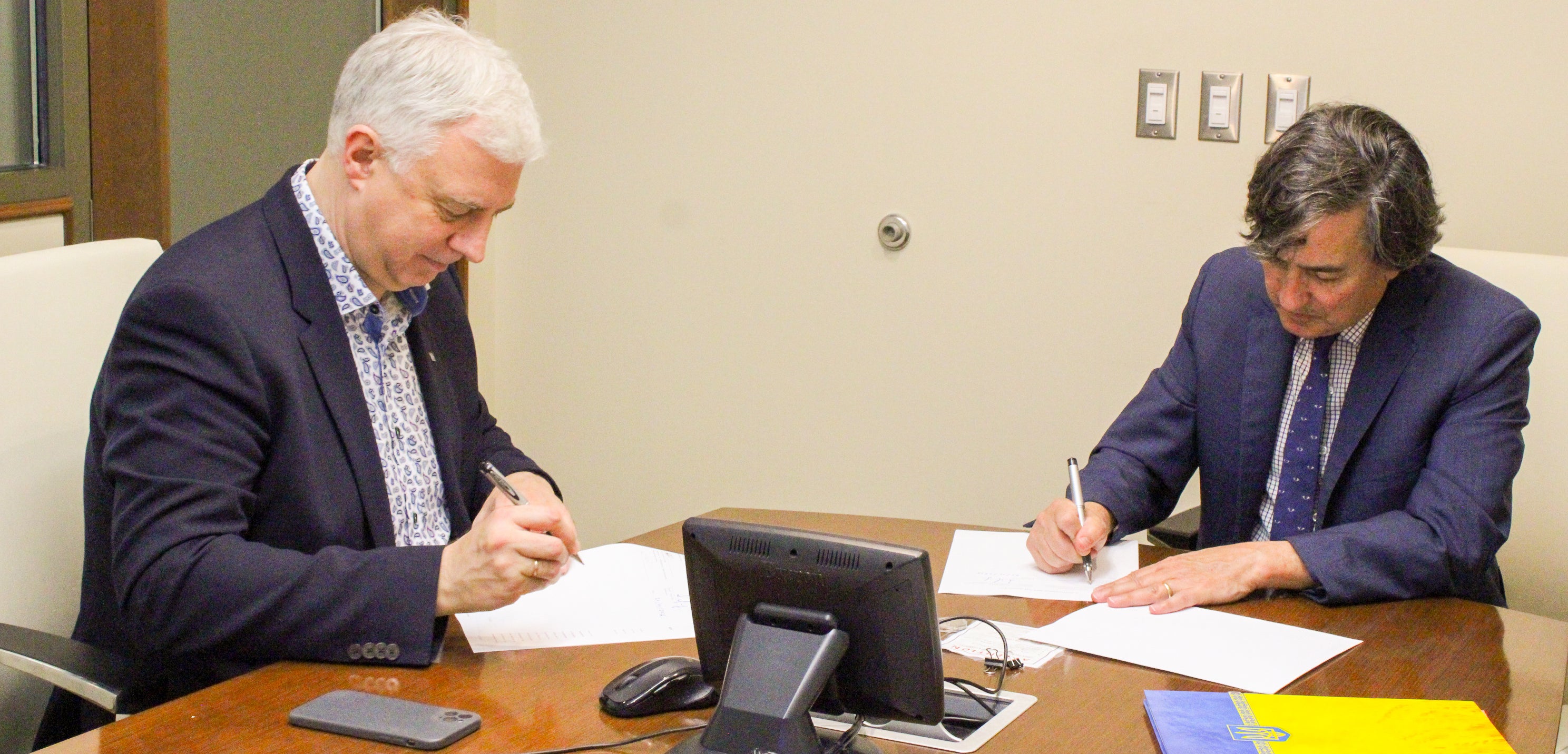 A signing ceremony with two men at a desk signing documents. 