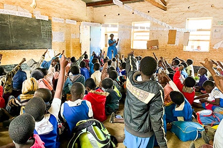 Students with their hands in the air and a teacher at the front of a classroom in Malawi.