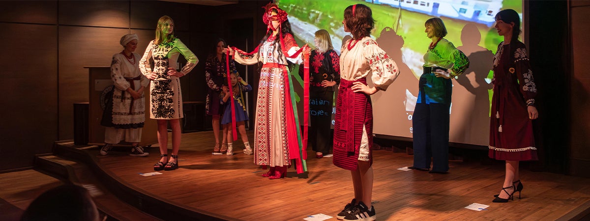A group of women on stage in front of a projection screen modeling vyshyvankas at a Vyshyvanka Day celebration.