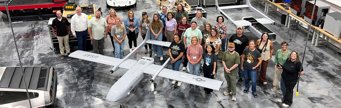 Group photo standing around an unmaned aircraft inside a warehouse.