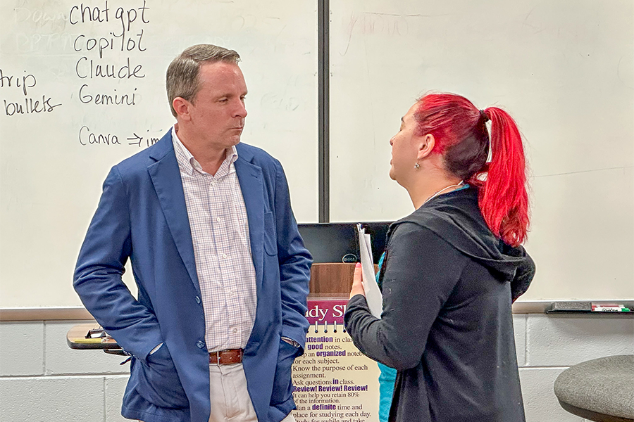 A person in a blue blazer conversing with another person who is holding a folder. They're in a classroom with a whiteboard featuring text mentioning "chatgpt," "copilot," "Claude," "Gemini," and "Canva" along with other notes. 