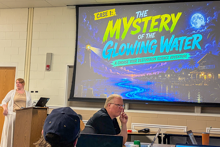 A classroom setting with a large screen displaying the title "Case 1: The Mystery of the Glowing Water" in a colorful design, featuring a moonlit scene of a lighthouse and water. A person stands near the screen, while another seated in front of a laptop appears to be listening.