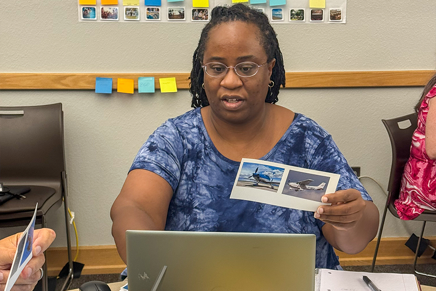 A person sitting at a table in front of a laptop, holding two airplane photos. Behind them is a wall with sticky notes and a small display of images.