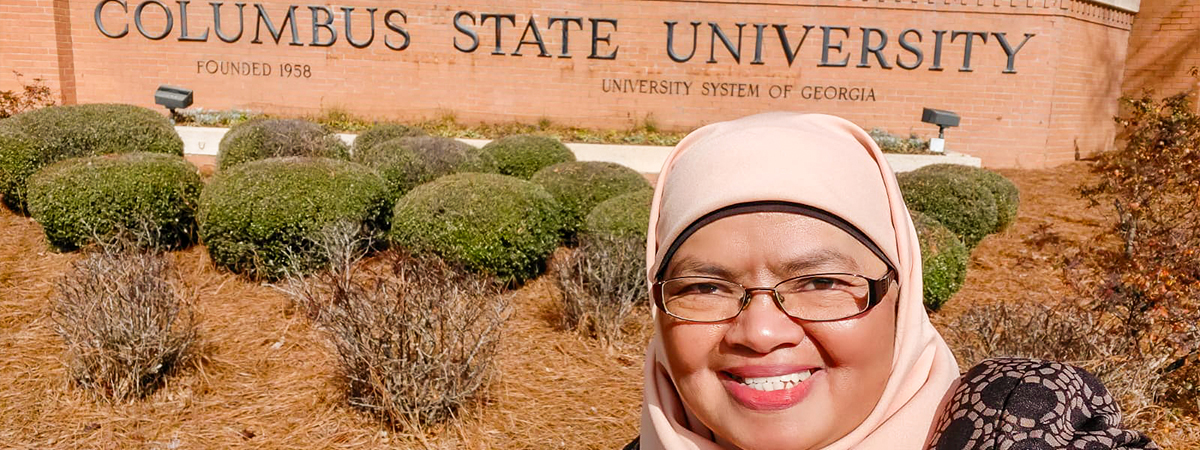 Dr. Ely selfie outside in front of a brick wall with a sign for Columbus State University.