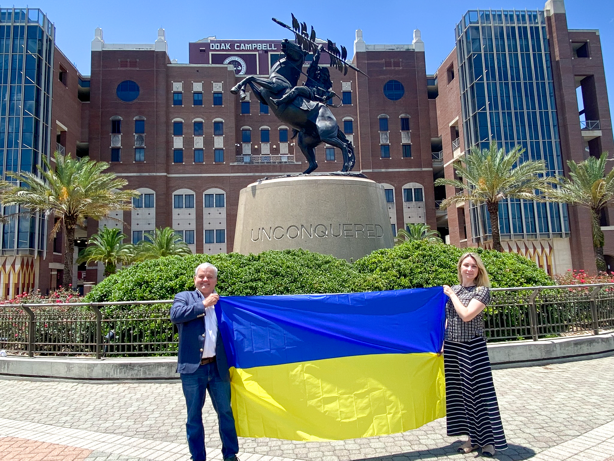 "Dr. Paziuk and Dr. Romanova pose with a Ukrainian flag in front of the Unconquered statute on the FSU campus."