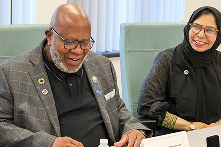 "President of the United Nations General Assembly, Mr. Dennis Francis sitting at a conference table smiling next to Communications Adviser to the President, Dr. Mariam Shaikh."