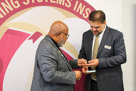 "President of the United Nations General Assembly, Mr. Dennis Francis presenting LSI Director Rabieh Razzouk with a UN pin in front of a large LSI globe logo. "