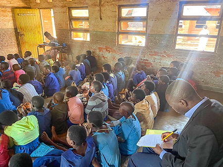 "This is a photo of a classroom with children sitting on the floor. There is a man in the background sitting in a chair taking notes. "