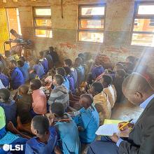 This is a photo of a classroom with children sitting on the floor. There is a man in the background sitting in a chair taking notes. 
