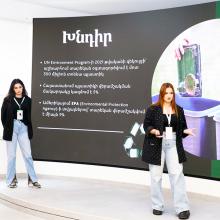 Two Armenian students pitching a business idea on stage in front of a large projection screen.