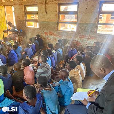 This is a photo of a classroom with children sitting on the floor. There is a man in the background sitting in a chair taking notes. 