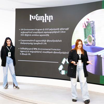 Two Armenian students pitching a business idea on stage in front of a large projection screen.