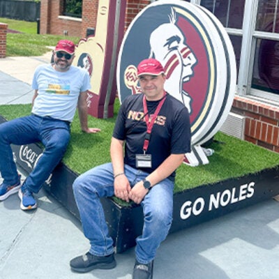 Dmytro Diadin, Ph.D. and Oleksandra Hrytstna, Ph.D. stiing at the FSU softball sdtadium in front of a large Seminole head FSU logo.