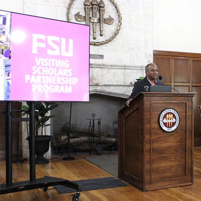 Participant in the Visiting Scholars Partnership Program addressing the room from a podium. There is a large screen TV next to him with the Visiting Scholars Partnership Program wordmark.