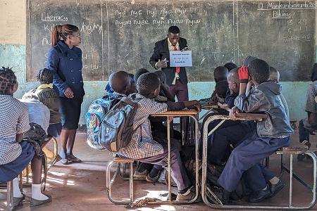 Man standing in front of a chalkboard at the head of a classroom of children. He is being observed by a woman while teaching.