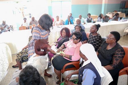 Group of women seated. One woman is standing with a book and looking at it with someone else. 