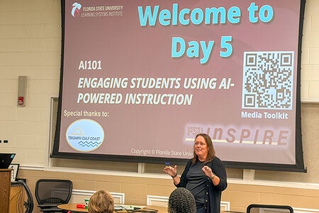 Anna Prindle is standing at the front of a classroom with a large projection screen behind her. 