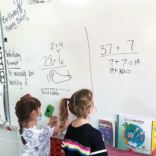 "Two children standing at a whiteboard working on math problems. "